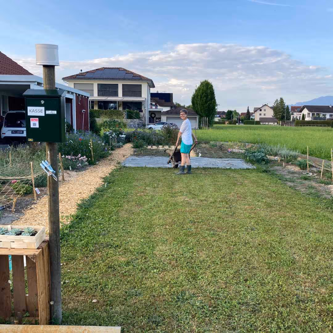 Caroline Ritter mit Schubkarre in einem gepflegten Garten mit Häusern und grünen Feldern im Hintergrund.