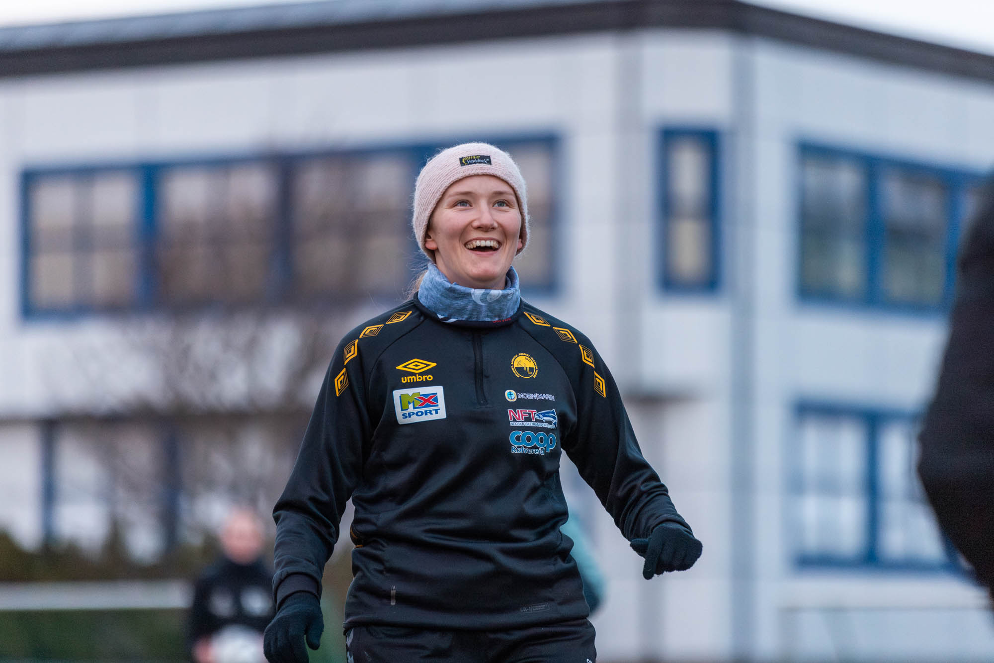 Smilende kvinne i sportsantrekk med lue og hansker utendørs med bygning i bakgrunnen.