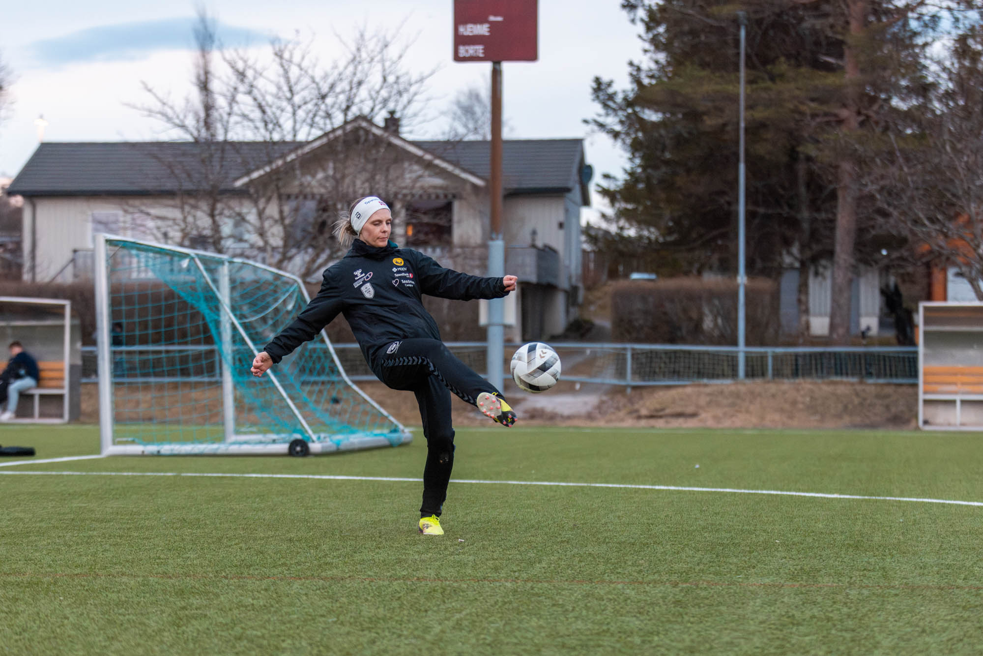 Person sparker en fotball på en fotballbane med mål og hus i bakgrunnen.