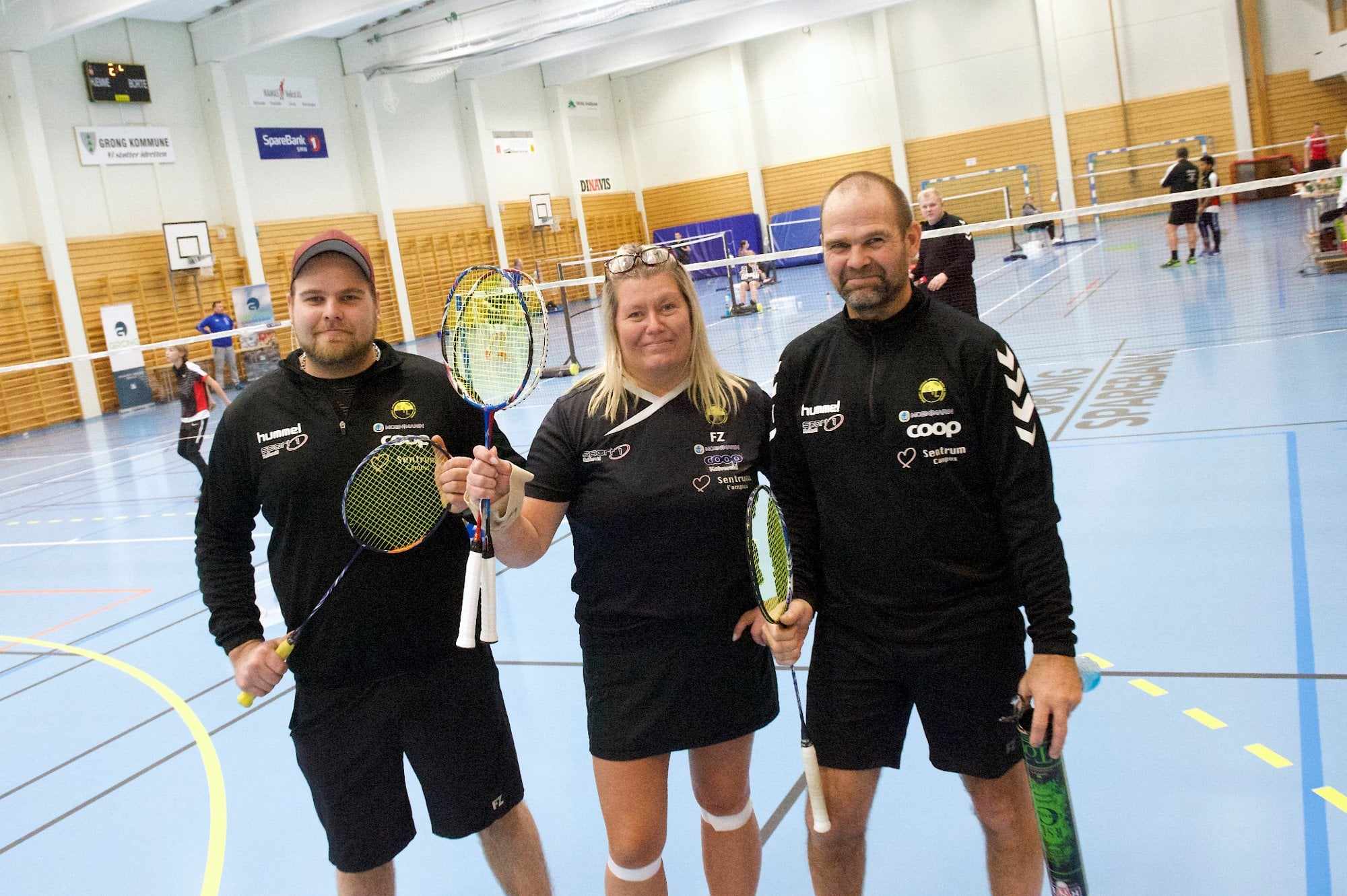Tre badmintonspillere i sportsklær holder racket og står i en badmintonhall.