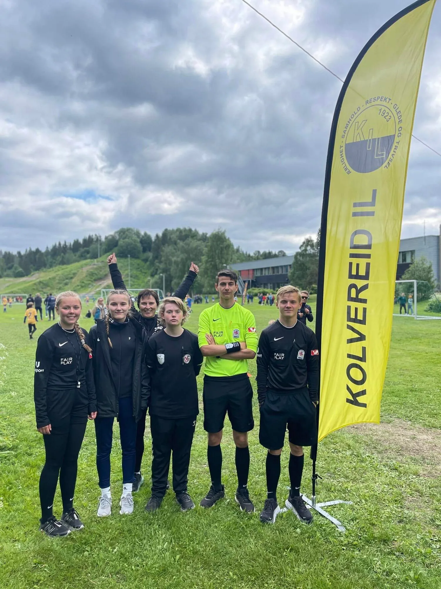 Seks personer, fem i svart og en i gult, står foran en gul Kolvereid IL-banner på en fotballbane.