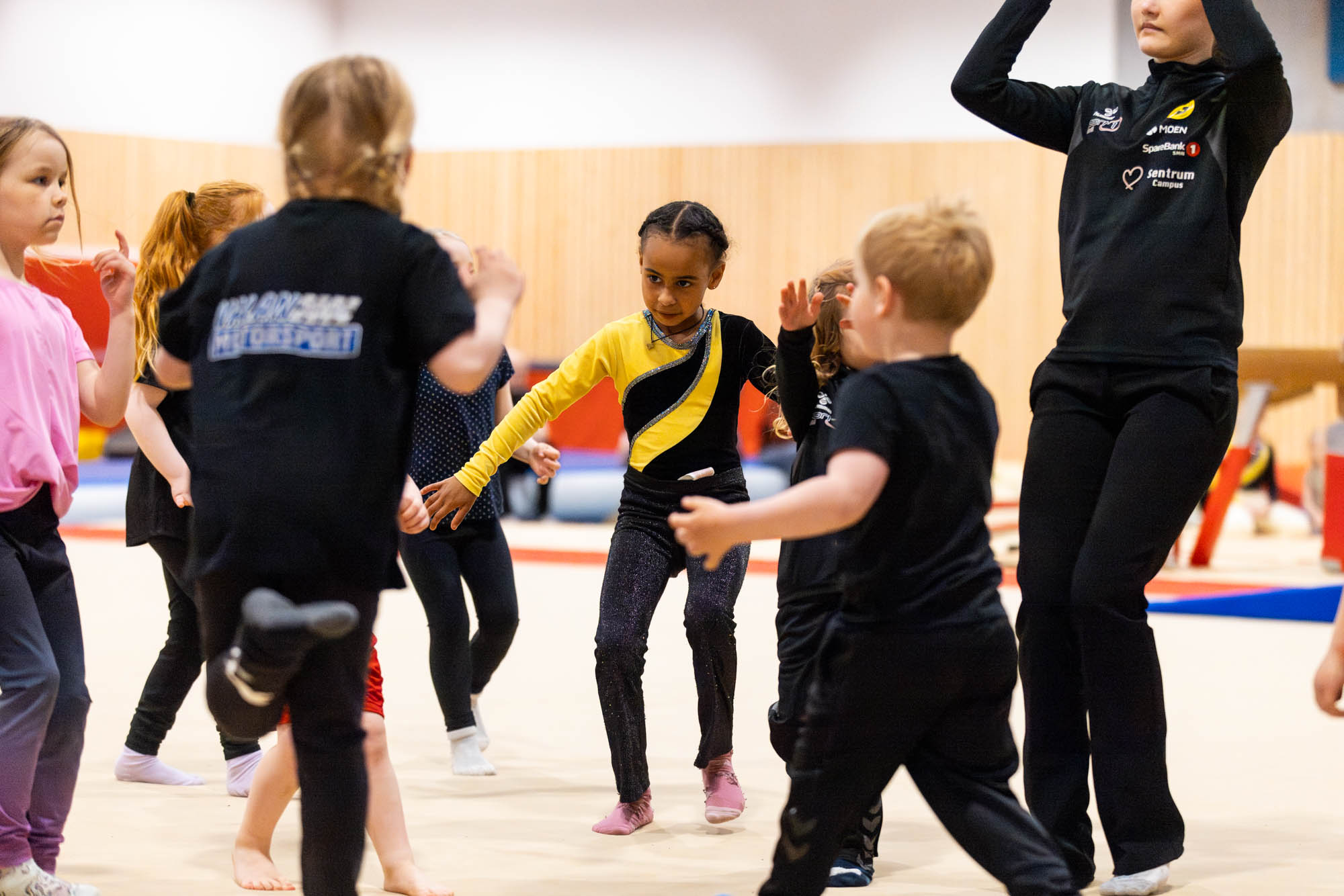 Barn trener gymnastikk i en gymsal mens en voksen instruktør veileder dem.