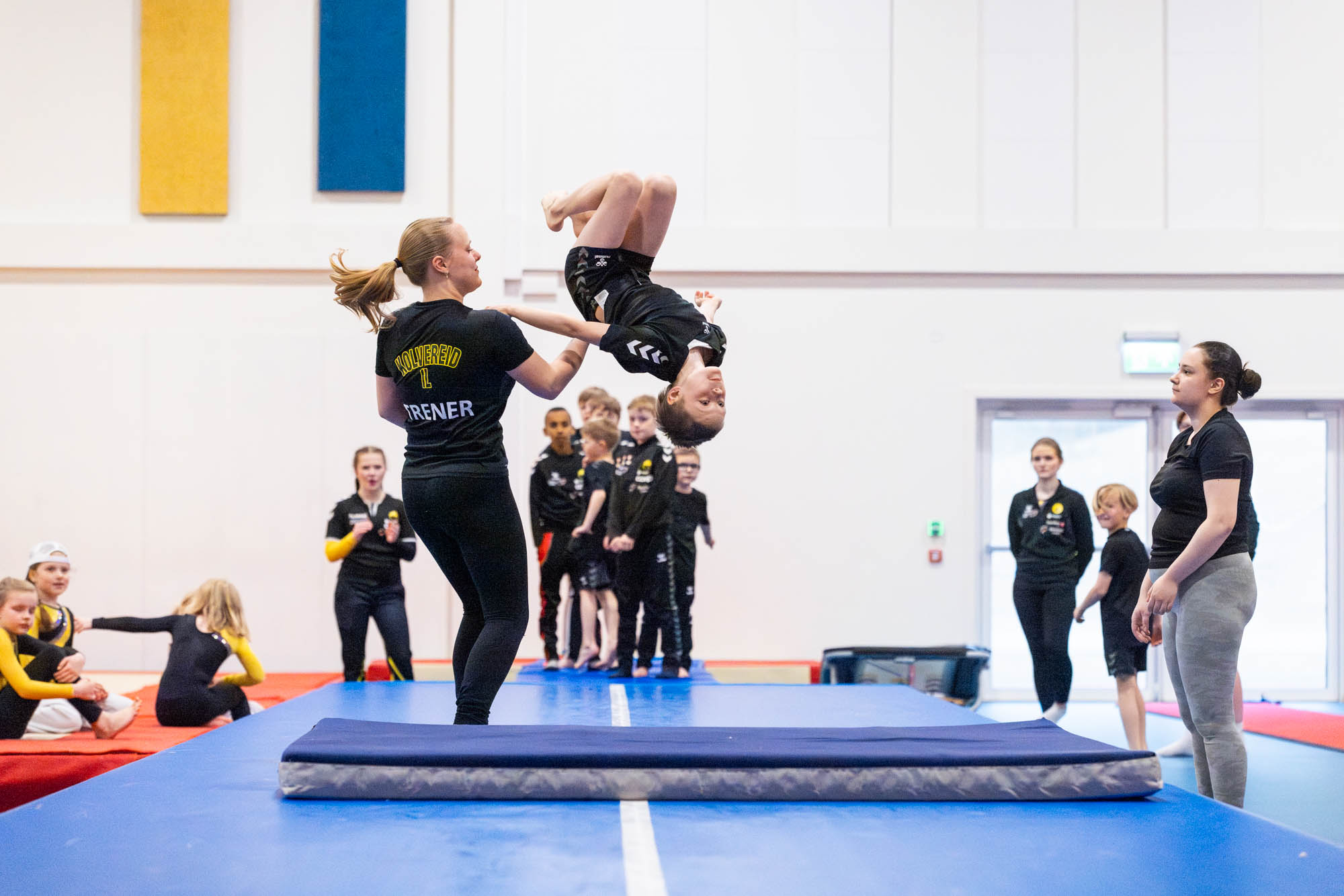 Barn utfører salto mens trener holder og flere barn følger med i en gymnastikksal.