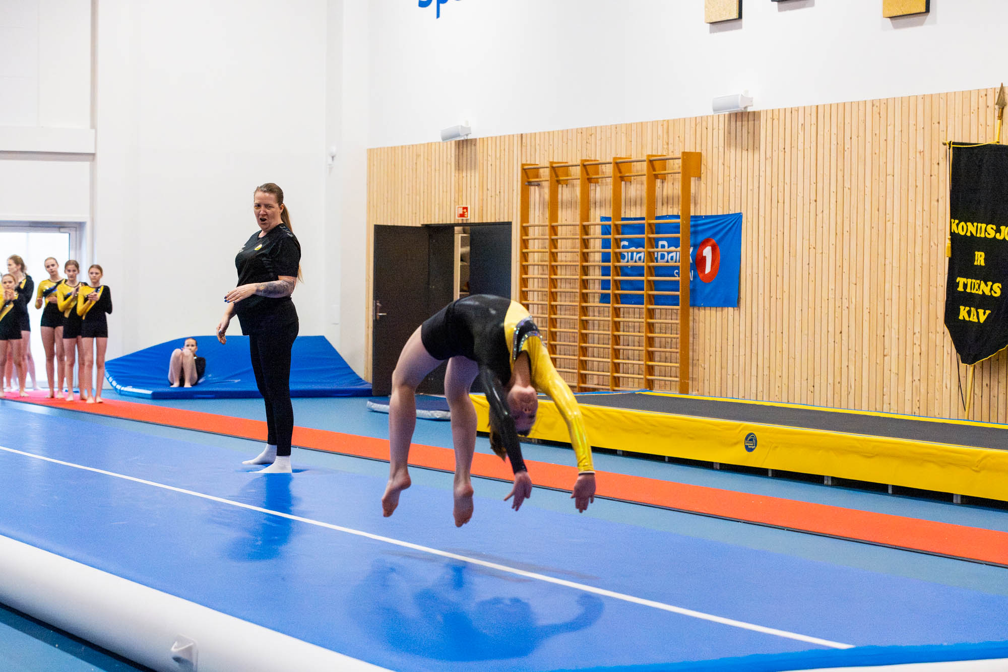 Ung gymnast som gjør en bakoverbøy på blå matter mens trener og lagkamerater følger med.