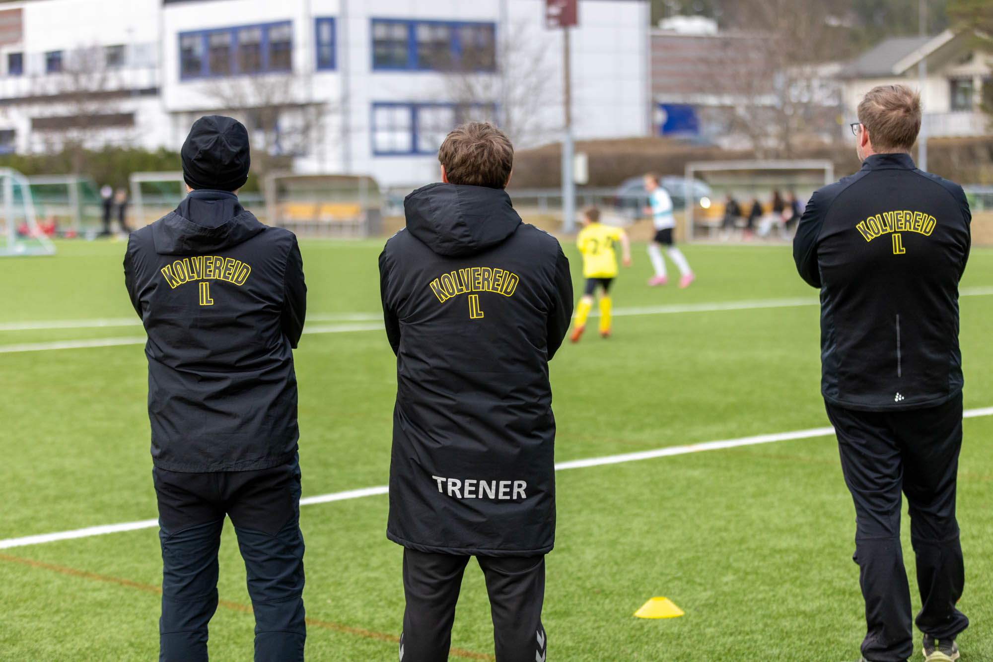 Three Kolvereid IL coaches in black jackets watch children playing soccer on a green field.