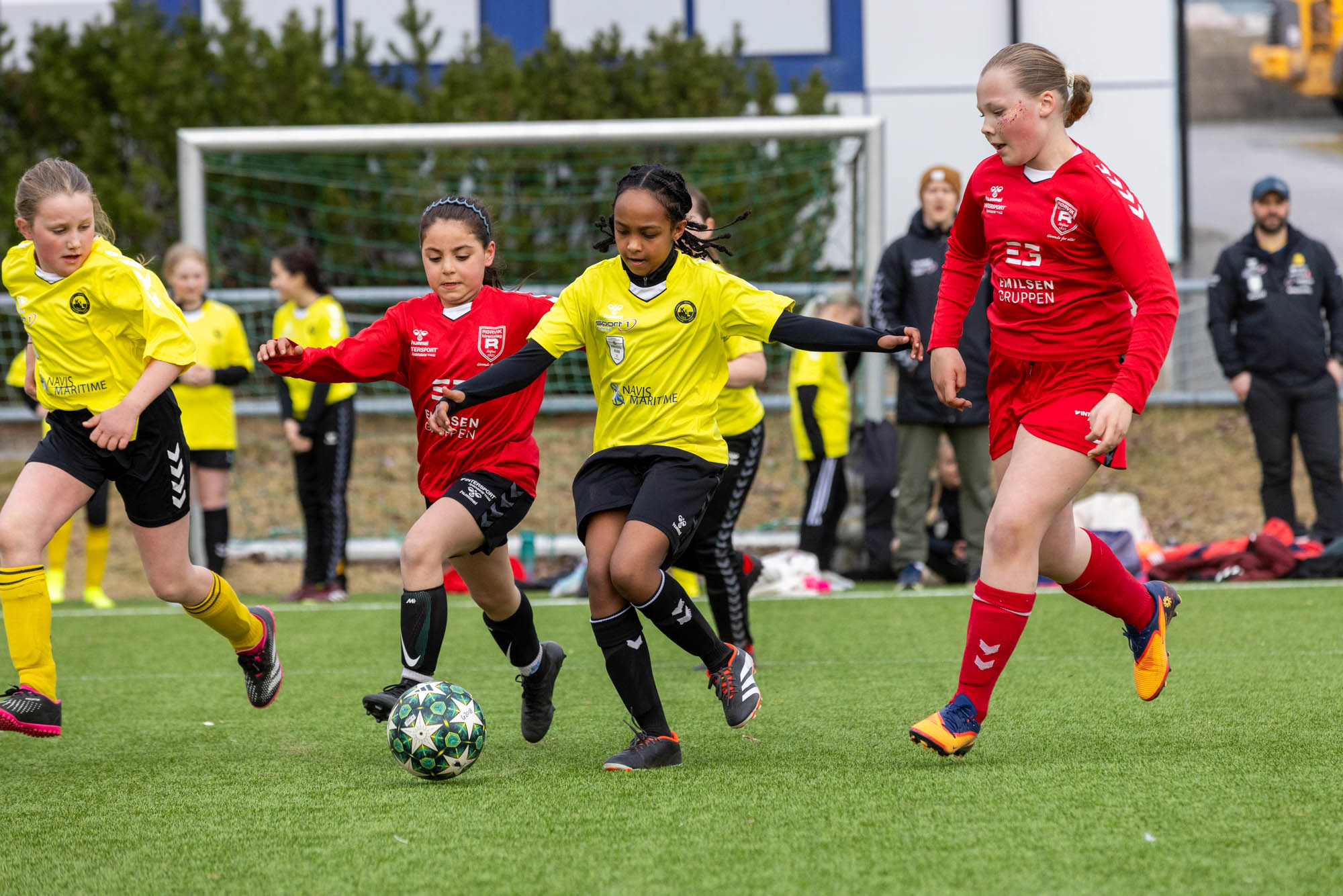 Barn spiller fotball, to i rødt og to i gult, konkurrerer om ballen på en grønn bane.