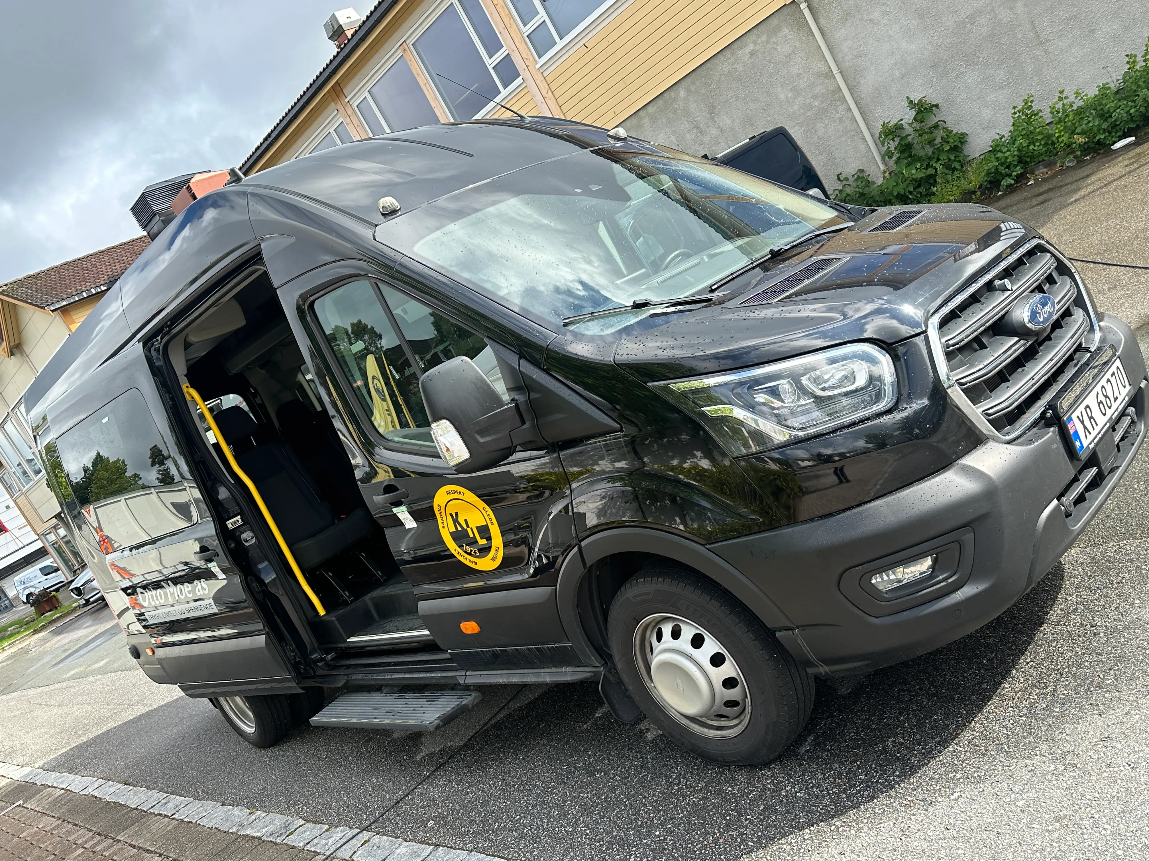 Svart minibuss med åpen side dør og gult KIL-logo på siden, parkert på en vei.