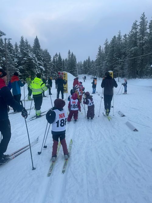 Barn står på ski i kø bak startlinjen i et snødekt terreng med tilskuere rundt.