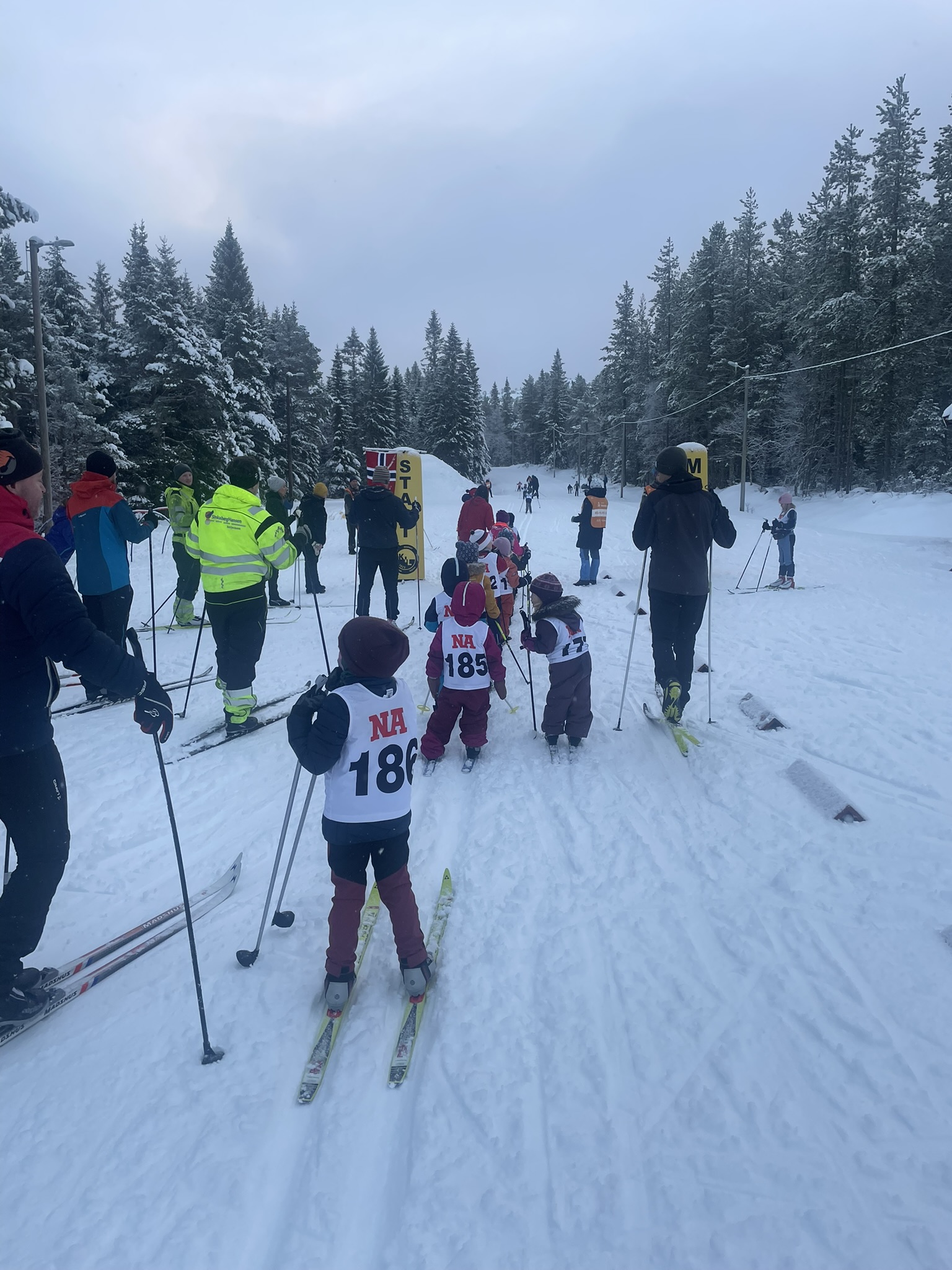 Barn står på ski i kø bak startlinjen i et snødekt terreng med tilskuere rundt.