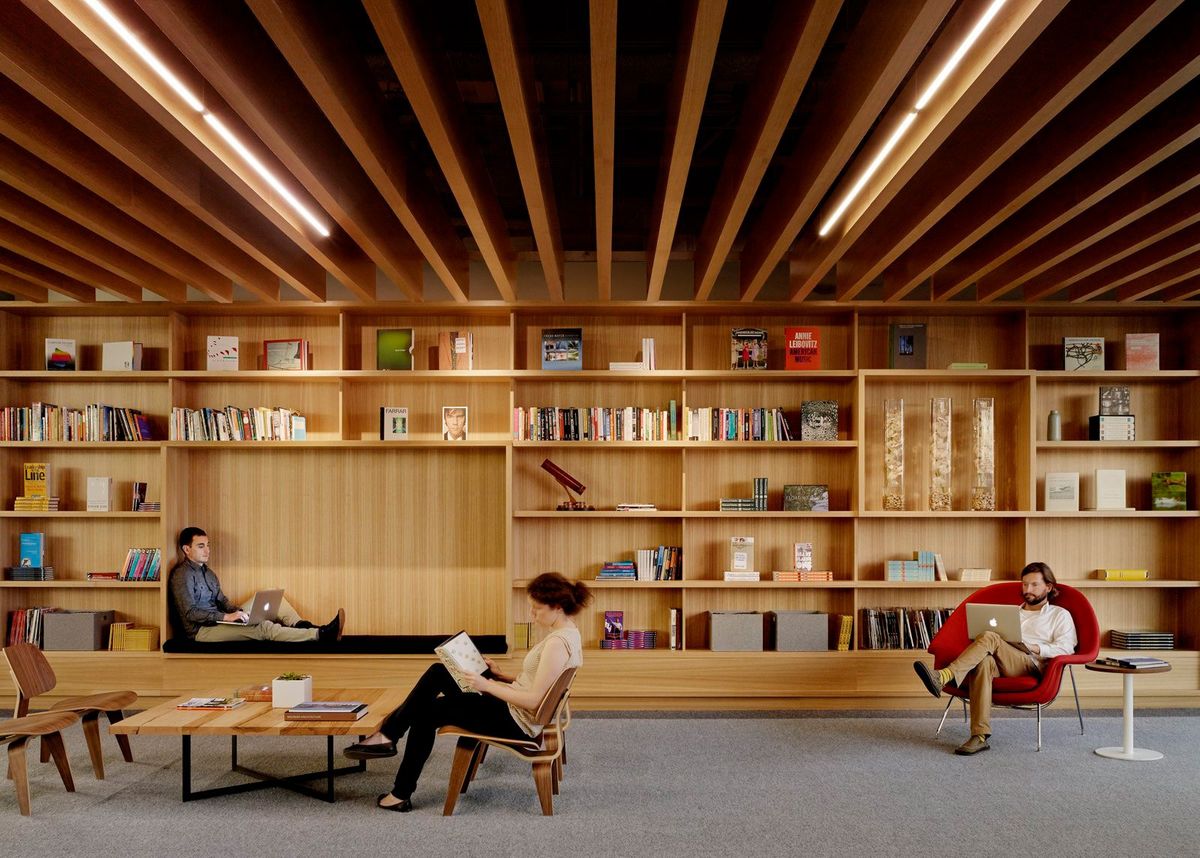 Modern library room with wooden shelves, three people working and reading, and wooden ceiling beams.