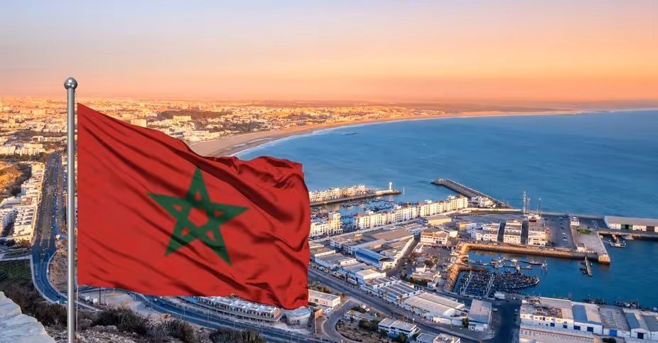 Red Moroccan flag with green star waving on a pole overlooking a coastal city and harbor at sunset.
