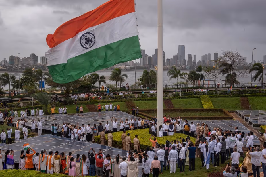 Large group of people gathered around a flagpole as the Indian national flag waves prominently against a cityscape background under a cloudy sky.