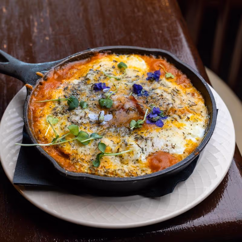 Baked dish with melted cheese, tomato sauce, herbs, microgreens, and purple edible flowers served in a cast iron skillet on a white plate.