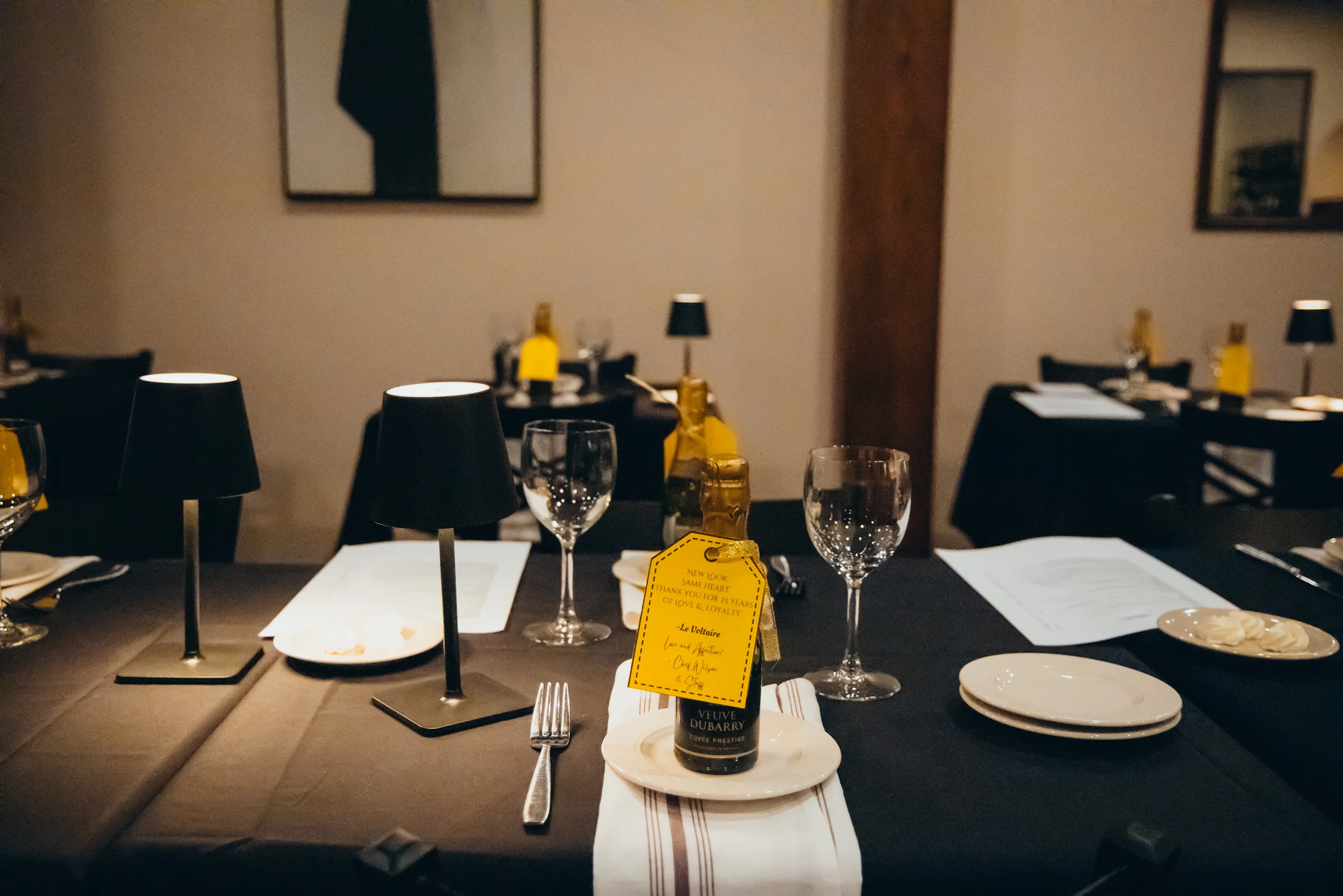 Table set for dining with black lamps, wine glasses, cutlery, small plates, and a bottle with a yellow tag in a cozy restaurant.