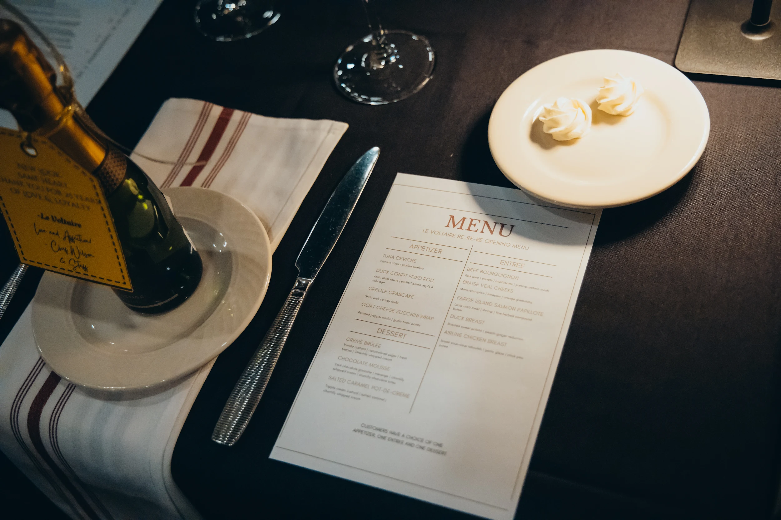 Table setting with a restaurant menu, butter plate with two butter curls, a knife, a bottle with a tag, and a striped napkin on a dark tablecloth.