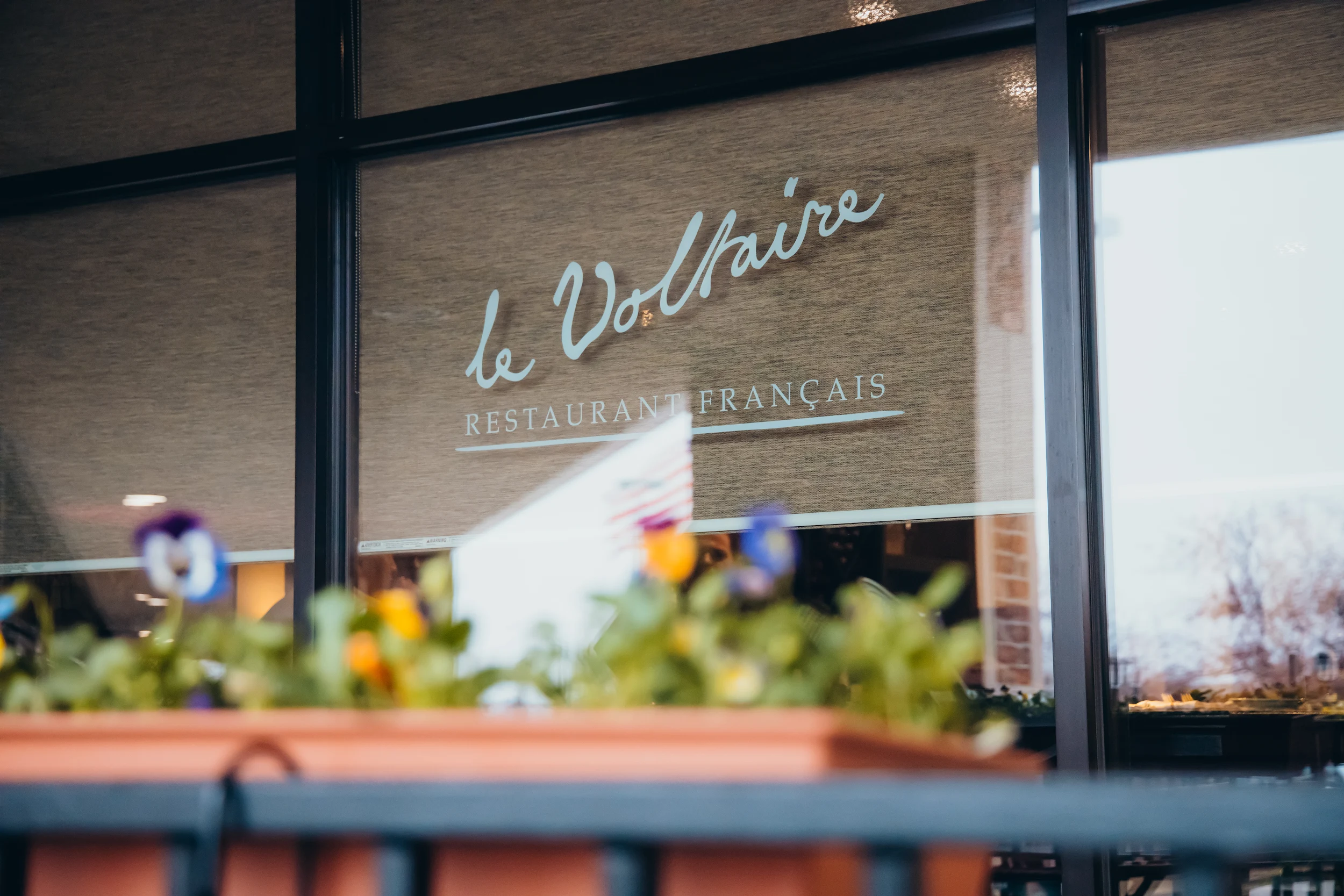 Window with the text 'le Voltaire RESTAURANT FRANÇAIS' partially covered by a flower box in front.
