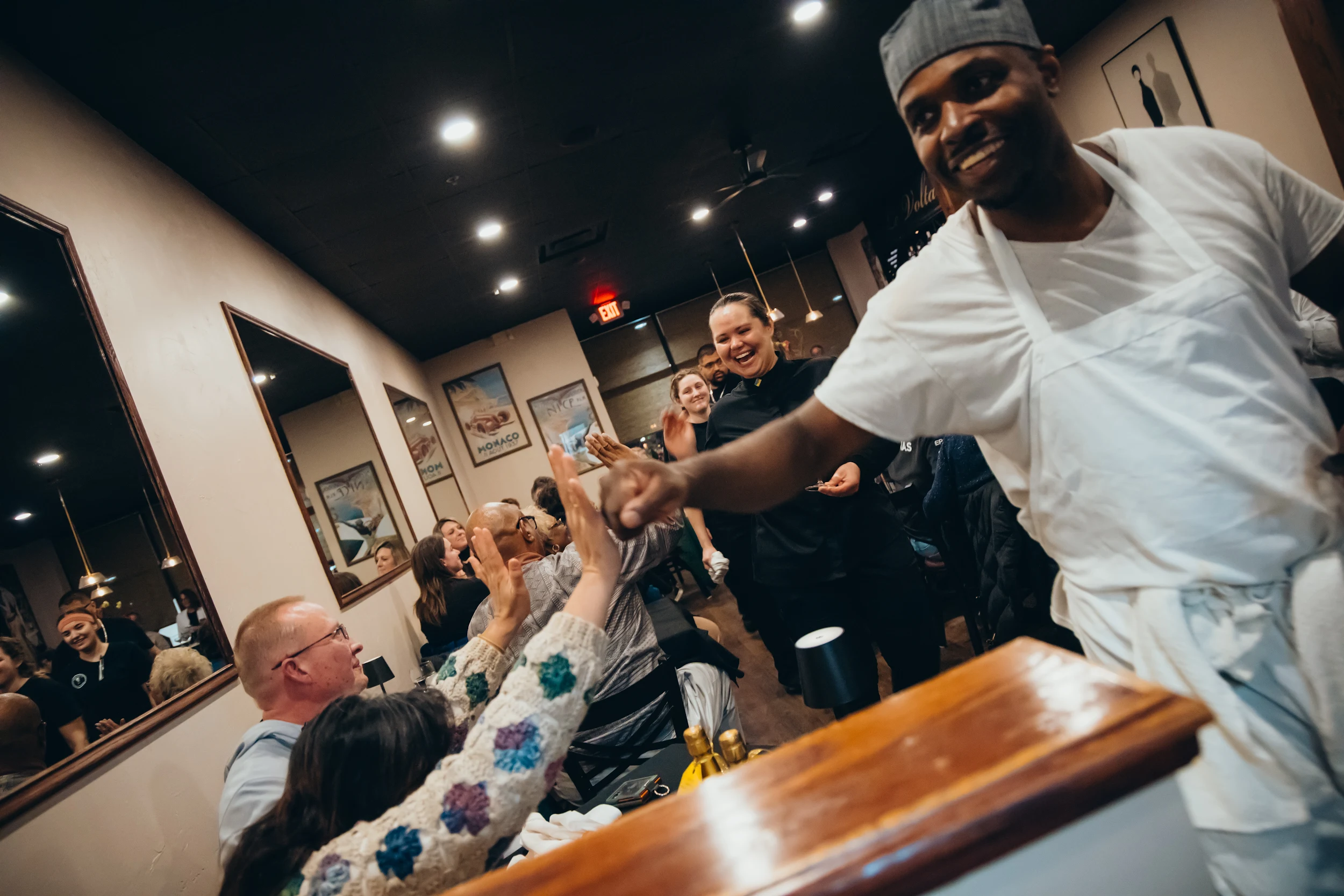 Chef in white apron high-fiving customers inside a lively restaurant.