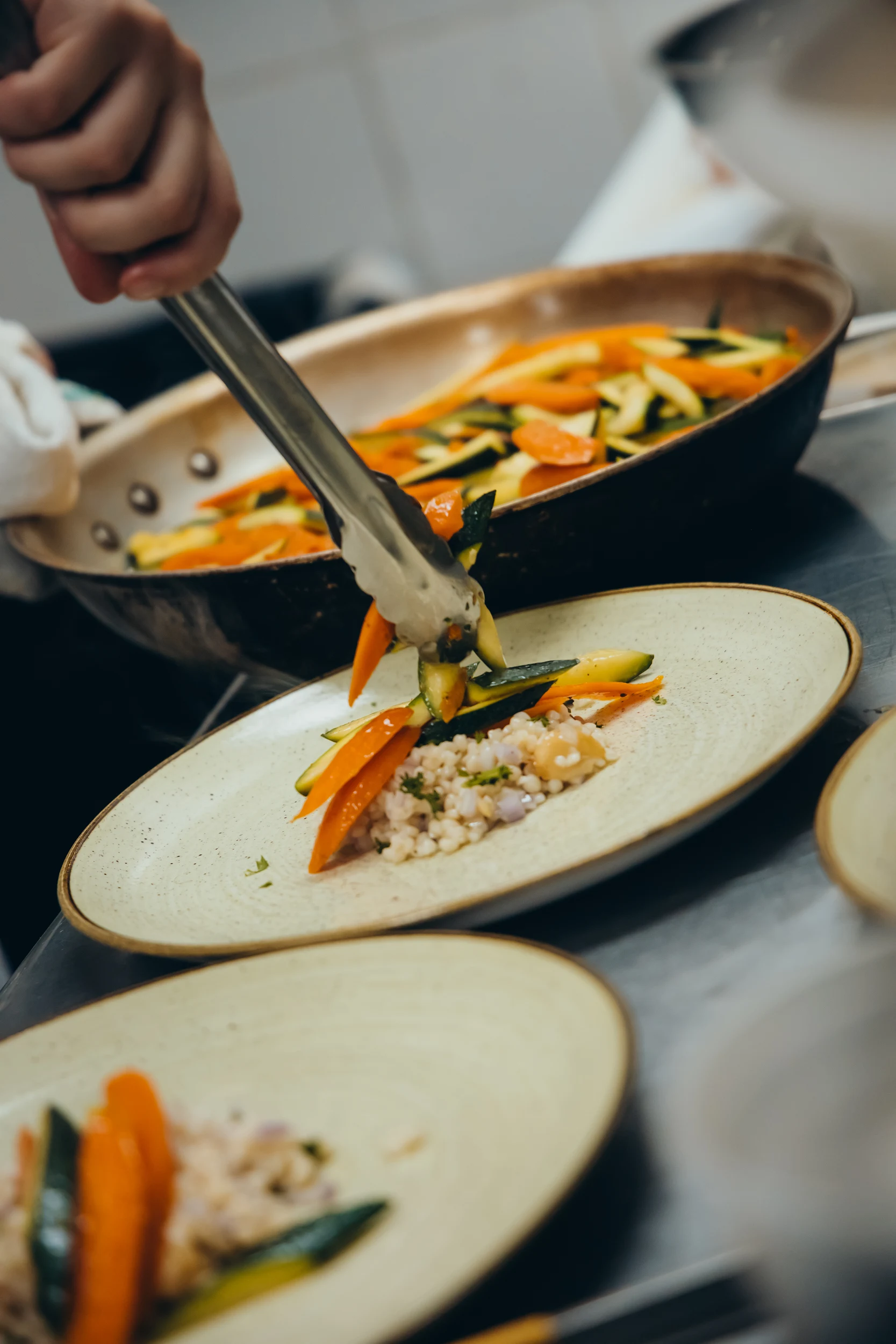 Hand using tongs to place cooked carrots and zucchini onto a plate with a grain salad.