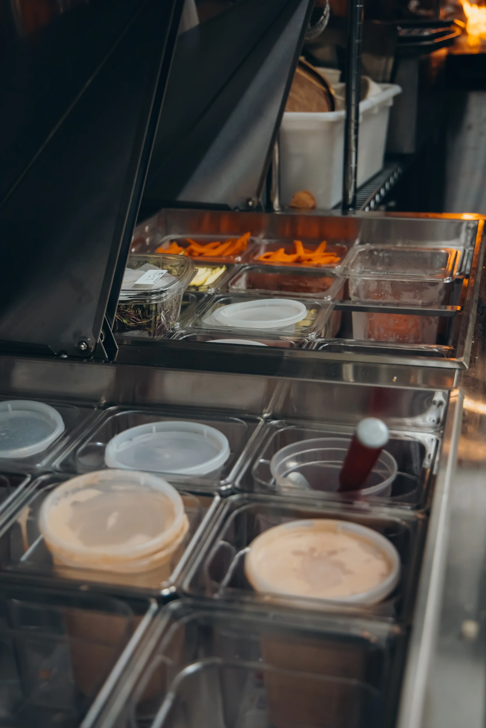 Close-up of a commercial kitchen prep station with various containers holding sauces, sliced carrots, and garnishes.