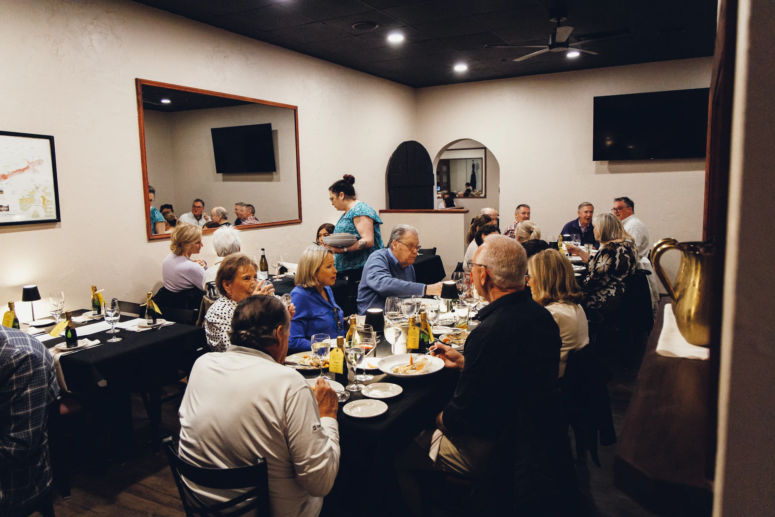 Group of people dining together in a warmly lit restaurant with black tablecloths and wine bottles on tables.