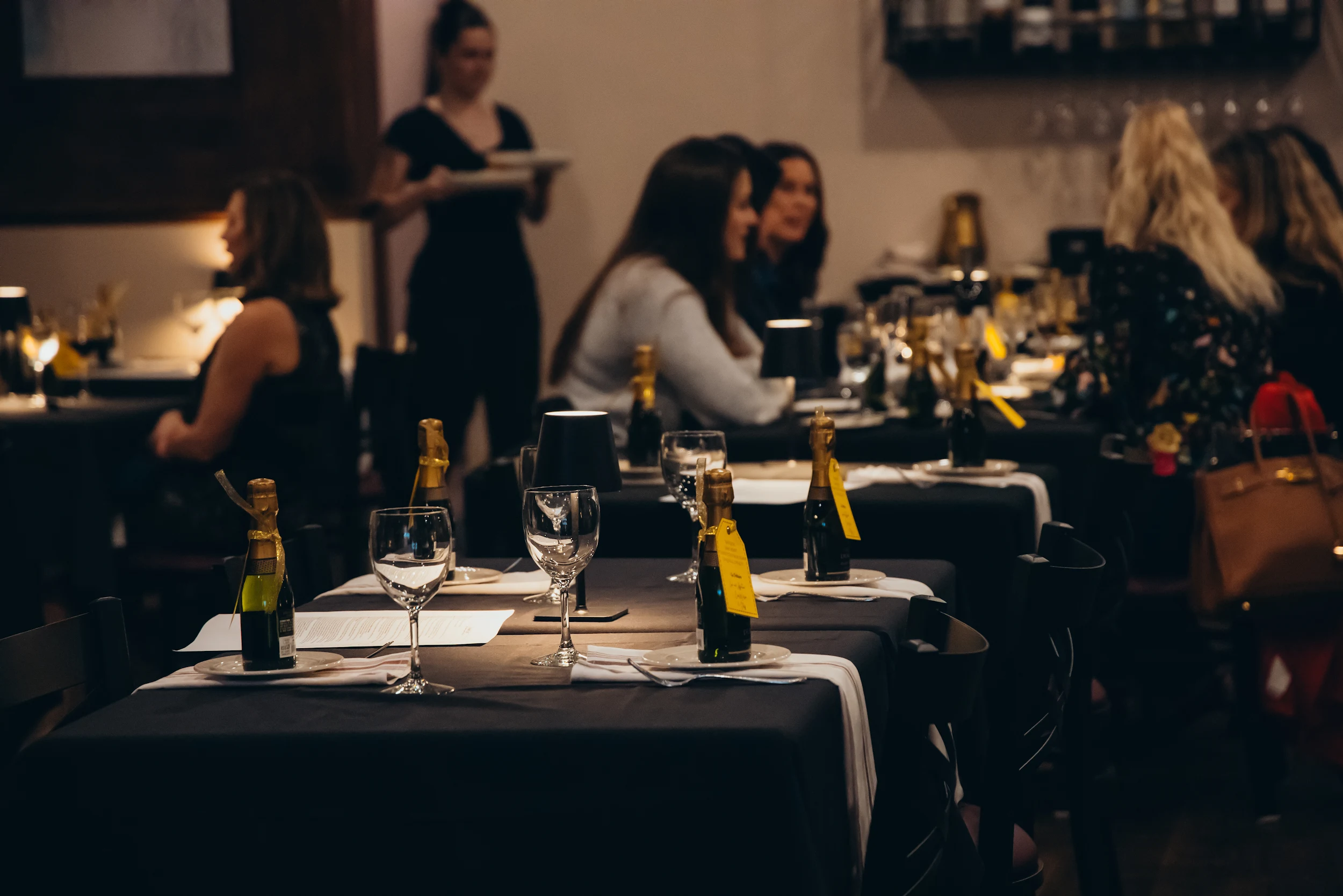 Dimly lit restaurant with tables set with wine glasses, mini champagne bottles, and black tablecloths, with diners and a server in the background.