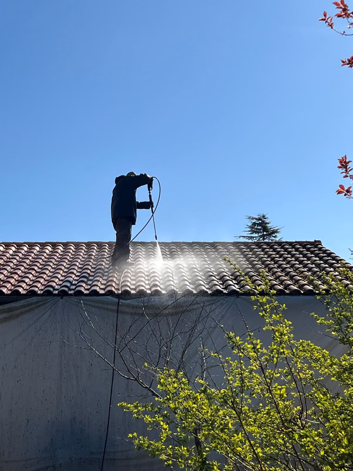 Un homme en équipement de sécurité nettoie un toit couvert de mousse avec un nettoyeur haute pression devant une maison résidentielle.