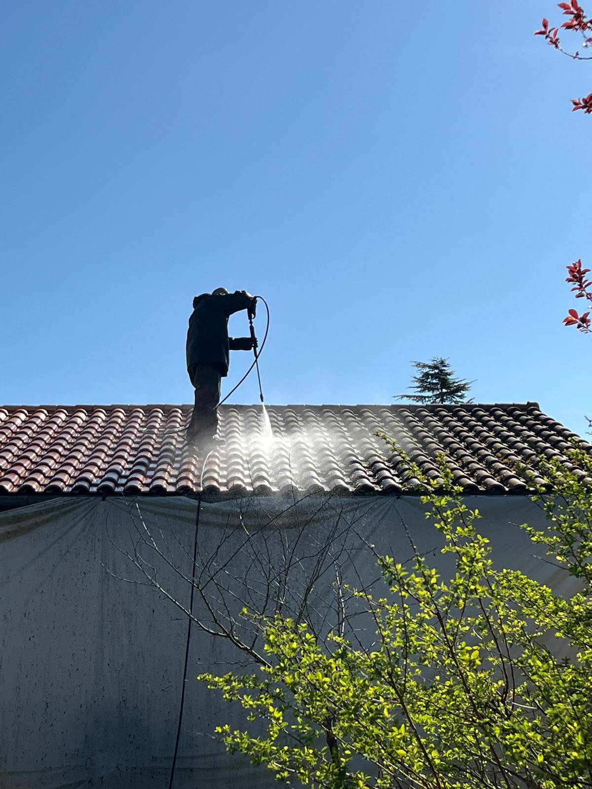 Un homme en équipement de sécurité nettoie un toit couvert de mousse avec un nettoyeur haute pression devant une maison résidentielle.
