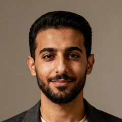 Portrait of a young man with short black hair, beard, and mustache, smiling slightly against a plain brown background.