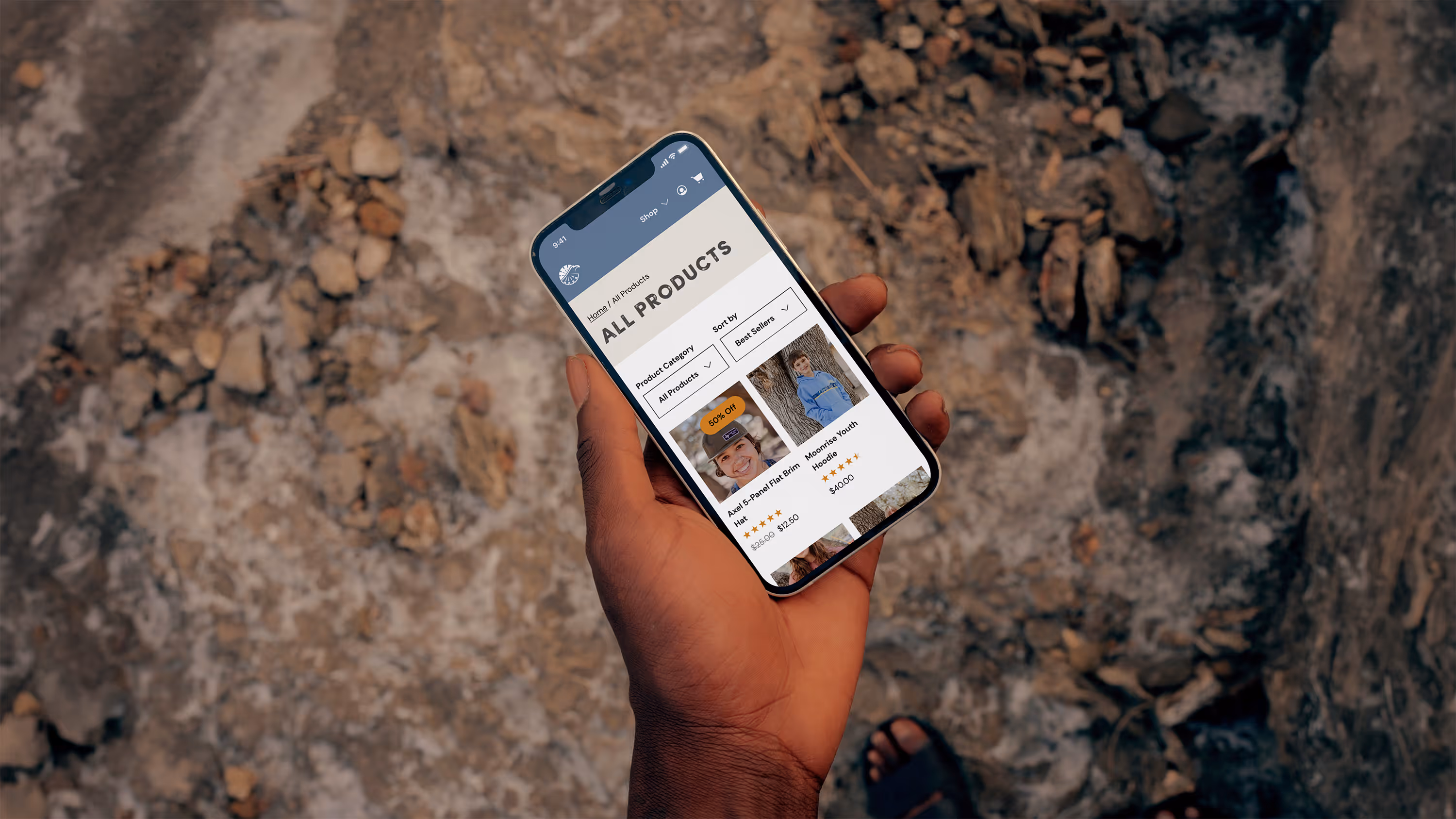 A person's hand holding a smart phone with Camp Eagle's ecommerce store open, with a rocky surface in the background.