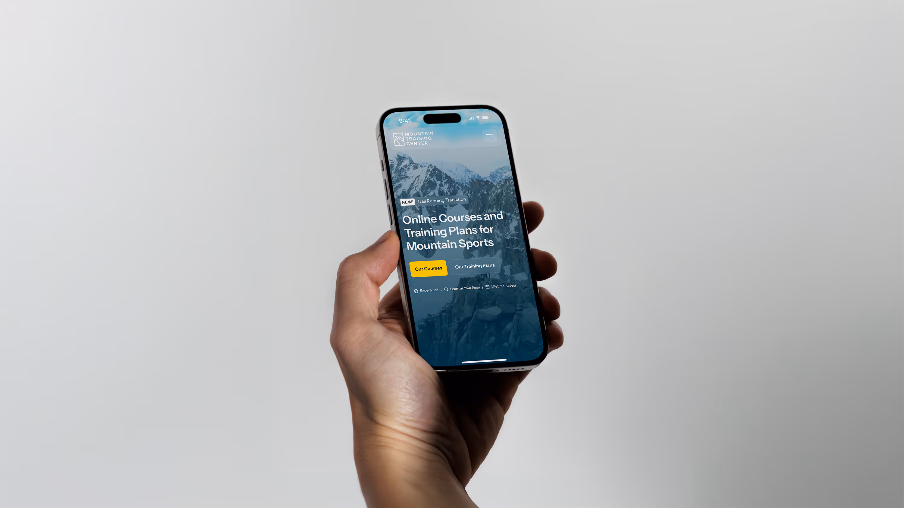 A hand holding a smart phone with the Mountain Training Center's home page hero section pulled up on it, with a white wall in the background.