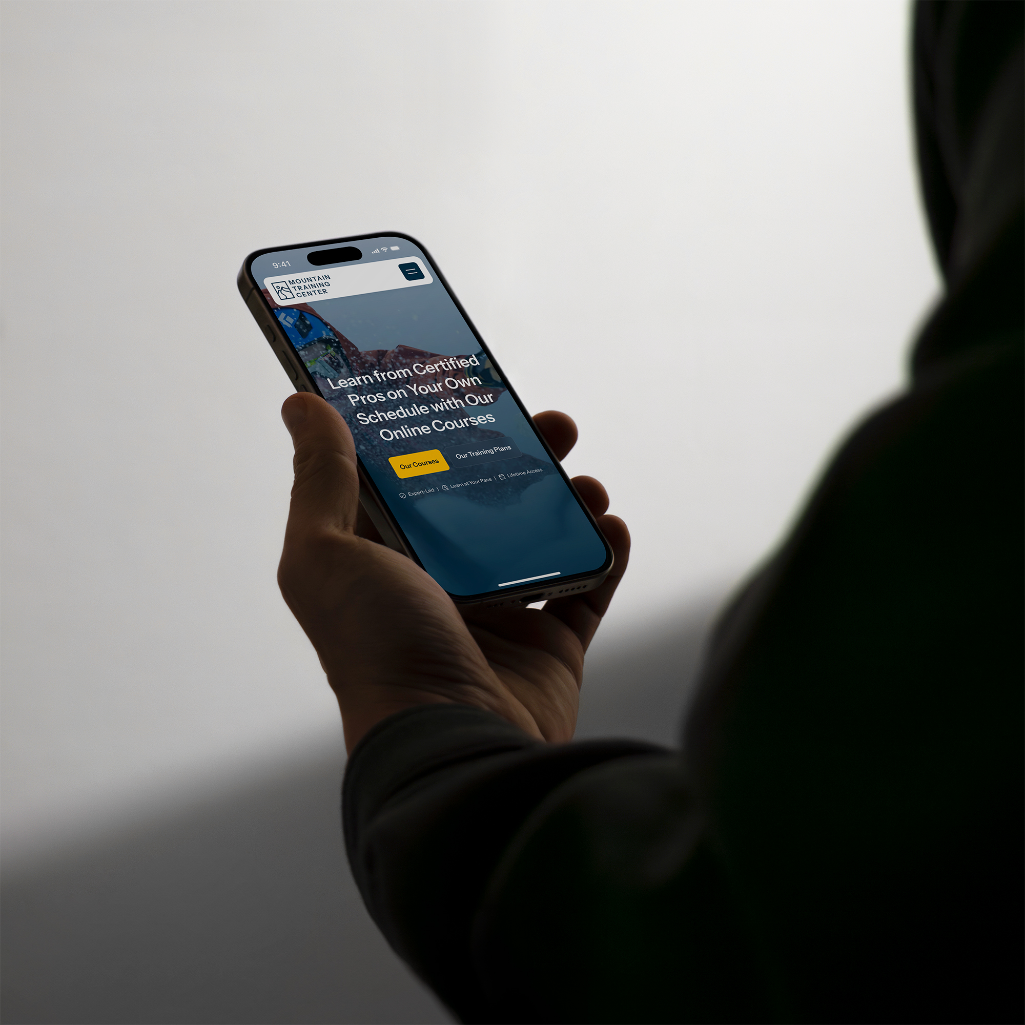 A hand holding a smart phone with the Mountain Training Center's home page call to action section pulled up on it, with a white wall in the background.