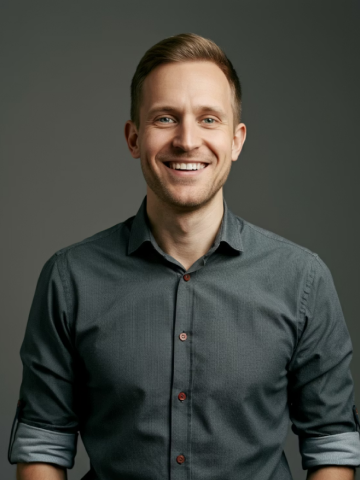 Smiling young man with short light brown hair wearing a dark grey button-up shirt with rolled-up sleeves.
