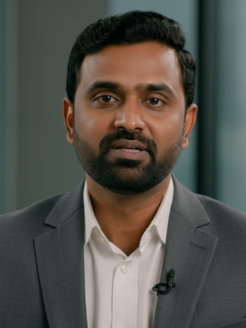 Portrait of a man with black hair and beard wearing a gray suit jacket and white shirt with a lapel microphone.