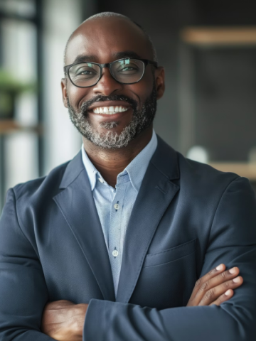 Smiling man wearing glasses and a blue blazer with arms crossed in a modern office.