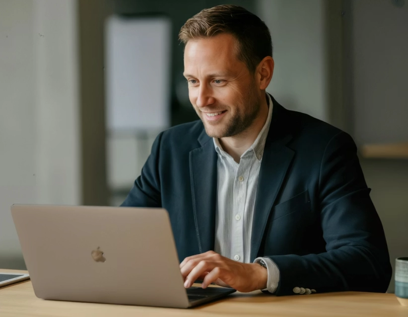 Smiling man in a blazer working on a laptop at a wooden table.