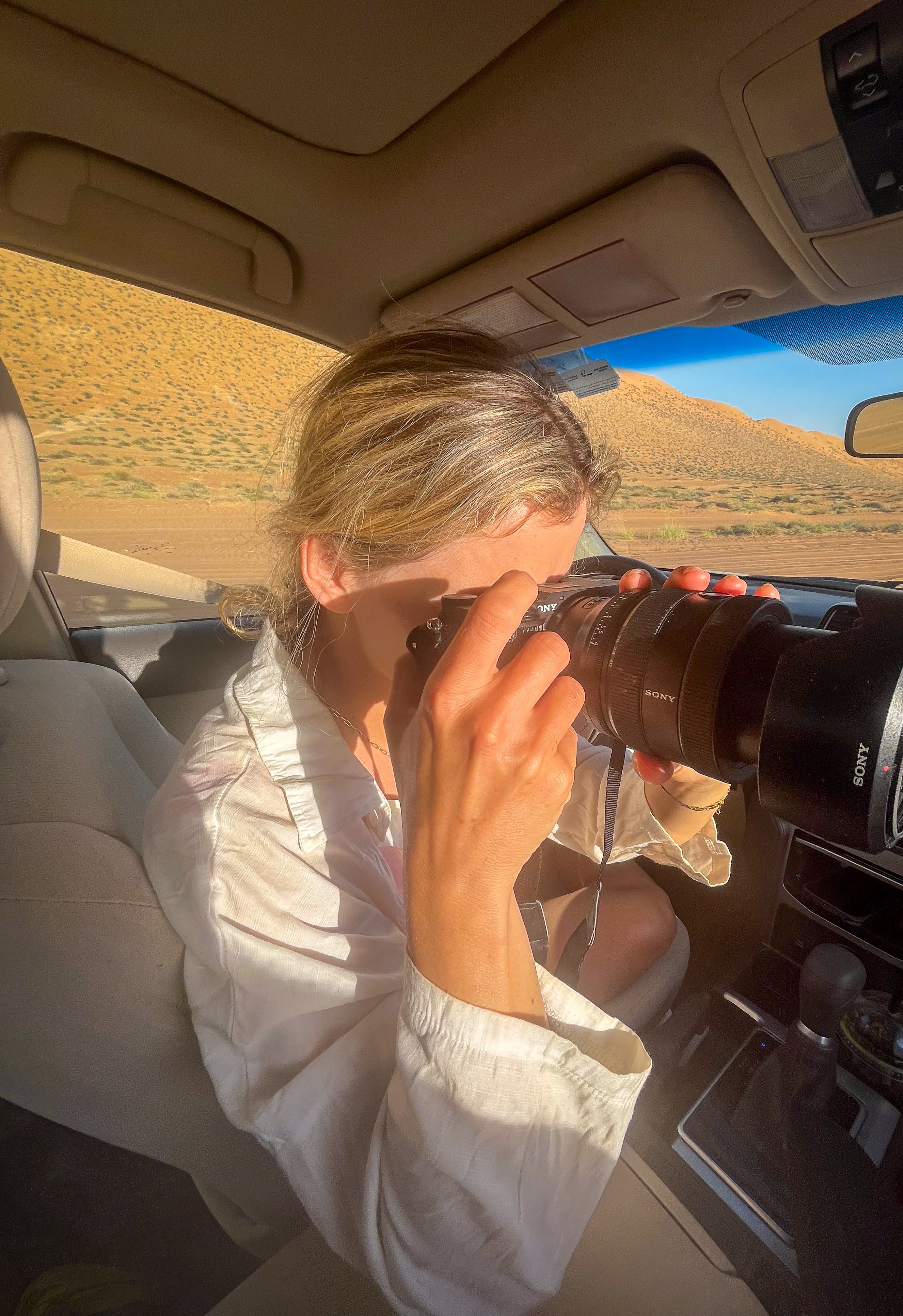 Me inside a car holding a Sony camera with a telephoto lens, taking a photo of a desert landscape outside.