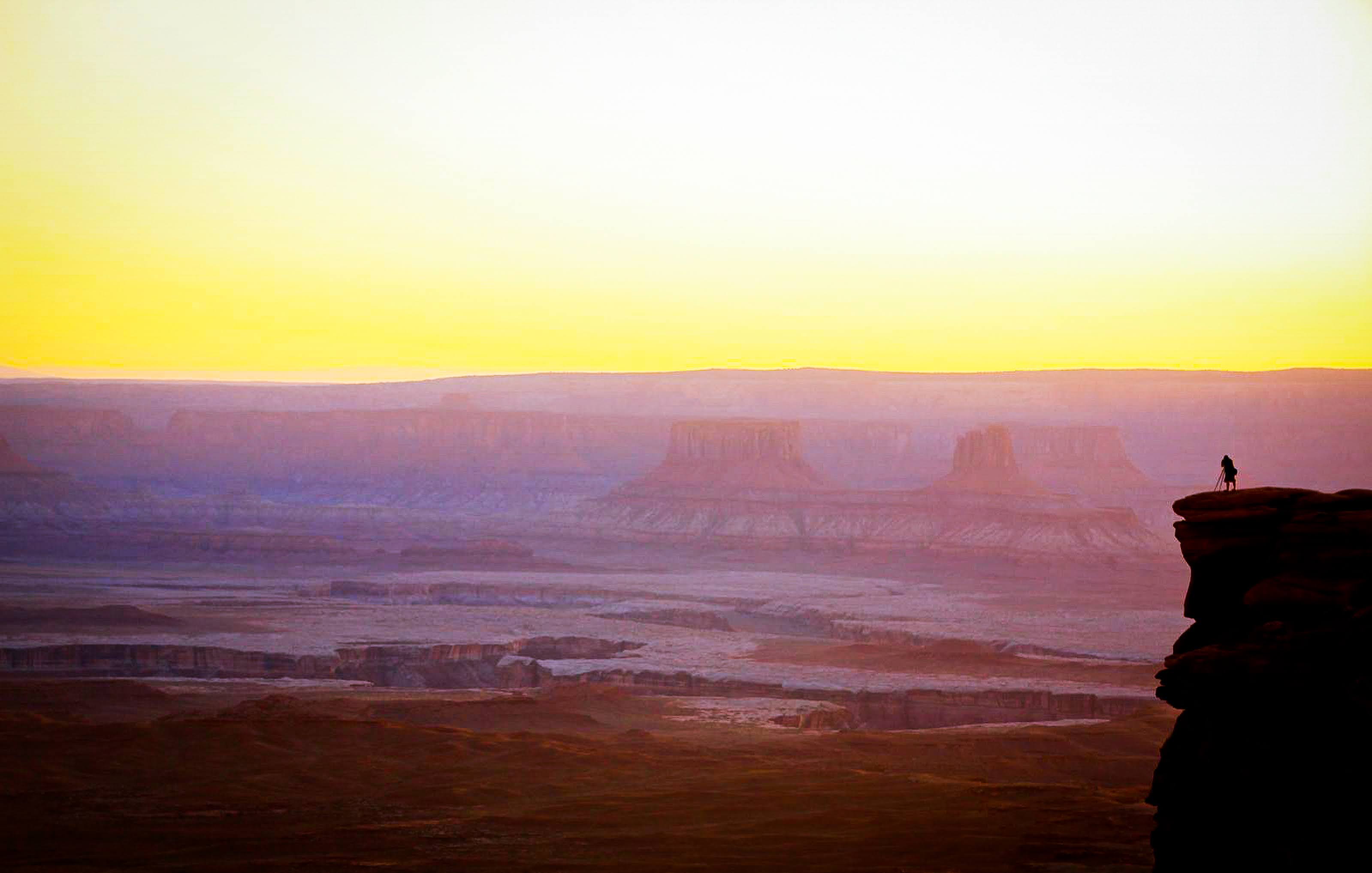 Silhouette of a person with a tripod on a cliff overlooking a vast canyon at sunset with a yellow and purple sky.