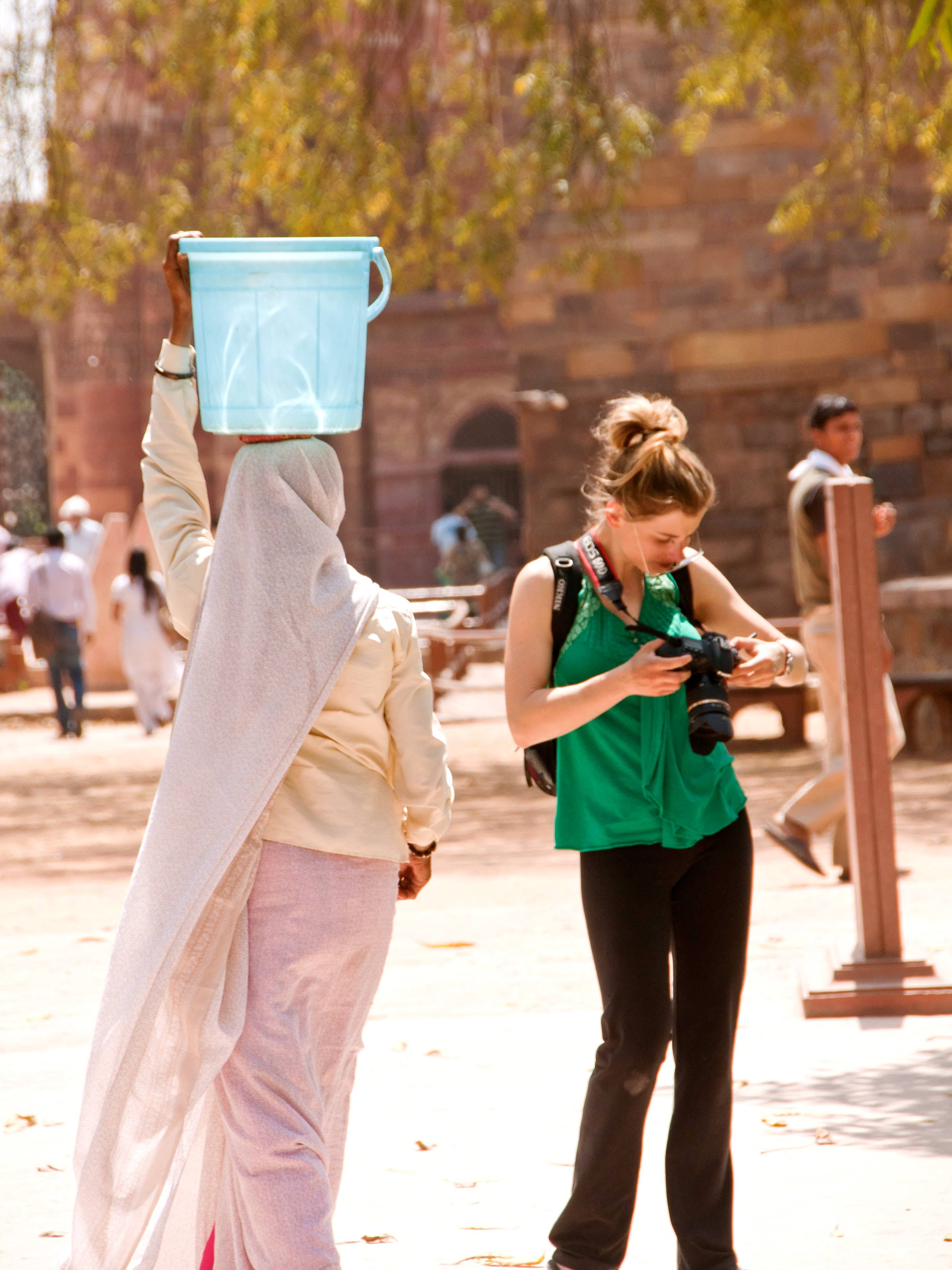 Yuna Rapoport in a blue bucket on her head walks past a female tourist in a green top who is looking at her camera outdoors.