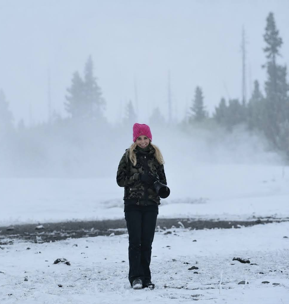 Yuna Rapoport in a pink knit hat and camouflage jacket holding a camera, standing on snowy ground with trees and mist in the background.