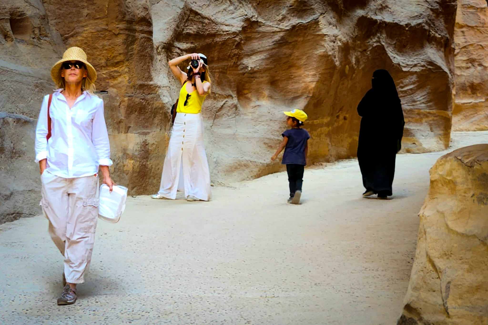 Four people in a rocky canyon; me, a woman in white and a straw hat walking, a woman in white pants and yellow top taking a photo, a child in a yellow cap walking, and a person in black traditional attire.