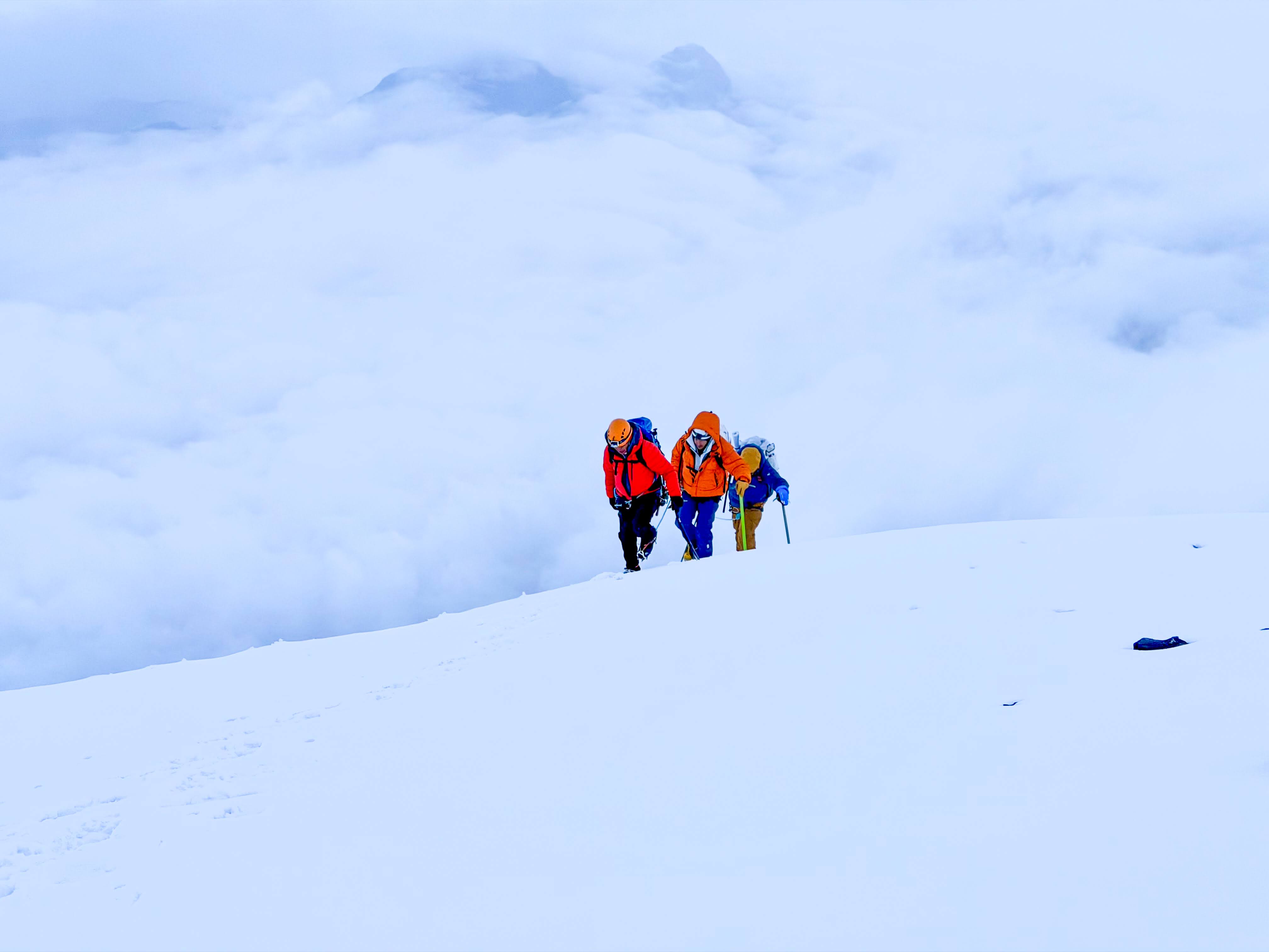Three climbers in bright jackets ascending a snowy mountain above the clouds.