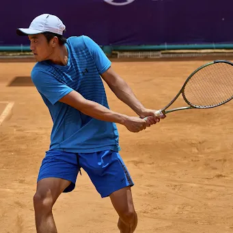Tennis player in blue shirt and shorts preparing to hit a backhand shot on a clay court.
