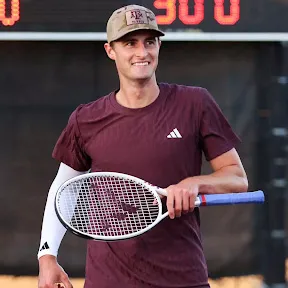 Mathis Ross, a smiling male tennis player in maroon shirt and beige cap holding a tennis racket on court.