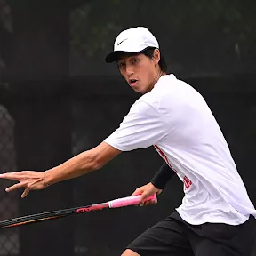 Erik Kuo, a man in white shirt and cap preparing to hit a tennis ball with a racket on a court.
