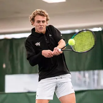 Harry Thursfield, a male tennis player in black shirt and white shorts hitting a backhand shot indoors.