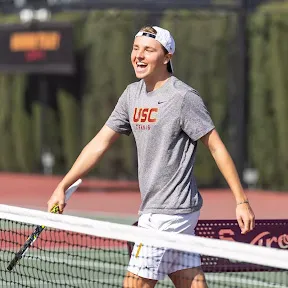 oscar weightman, a young male tennis player in USC shirt and white cap smiling on a tennis court near the net.