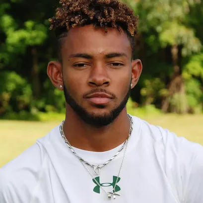 noah williamson, a young man with short curly hair and beard wearing a white Under Armour shirt and silver necklaces, outdoors with greenery in the background.