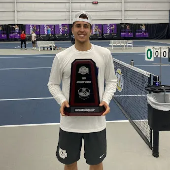 a young male athlete in a white cap and shirt holding a tennis championship trophy on an indoor tennis court.