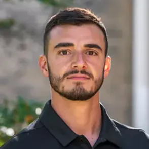 Portrait of a young man with short dark hair, beard, and mustache wearing a black collared shirt, with a blurred outdoor background.