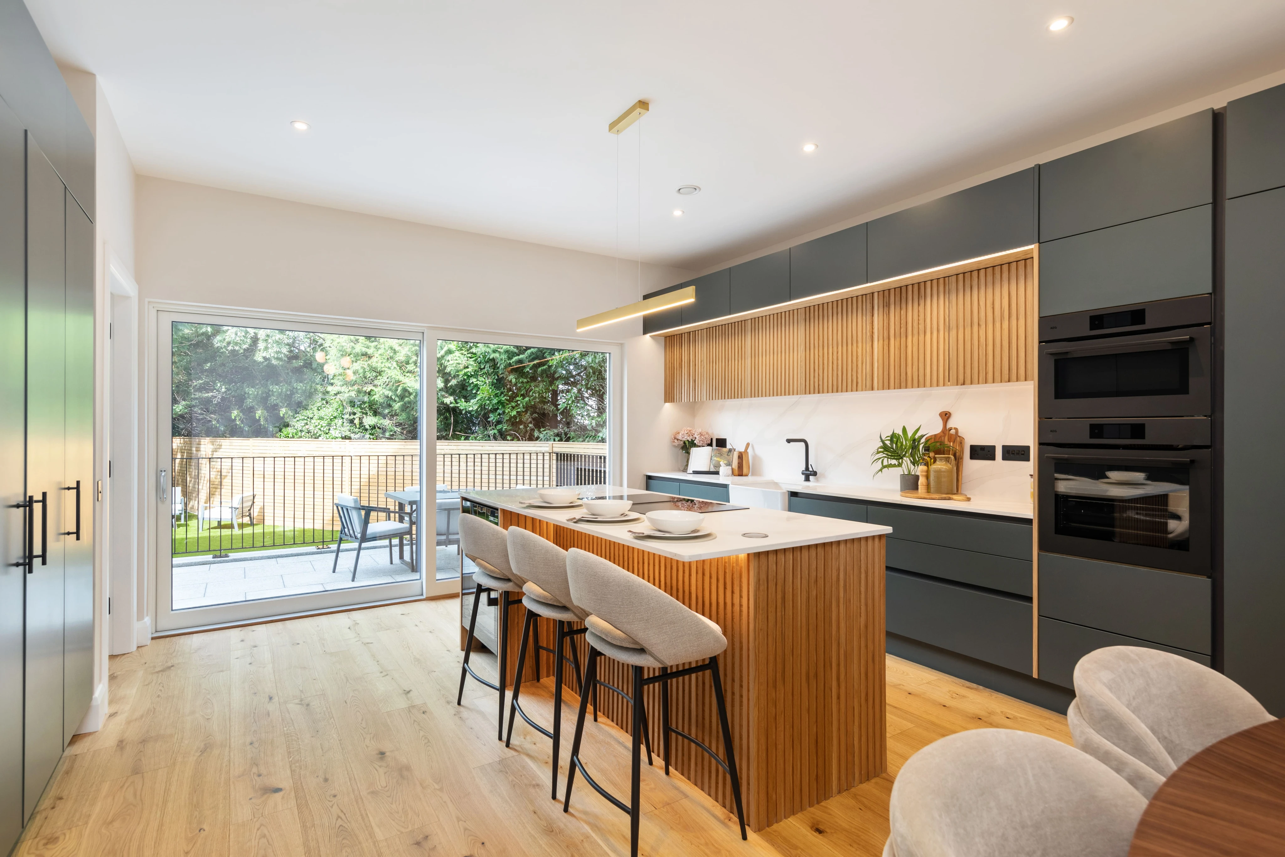 Modern kitchen with wooden floor, central island with seating, dark grey cabinets, and large sliding glass doors opening to a patio with outdoor furniture.